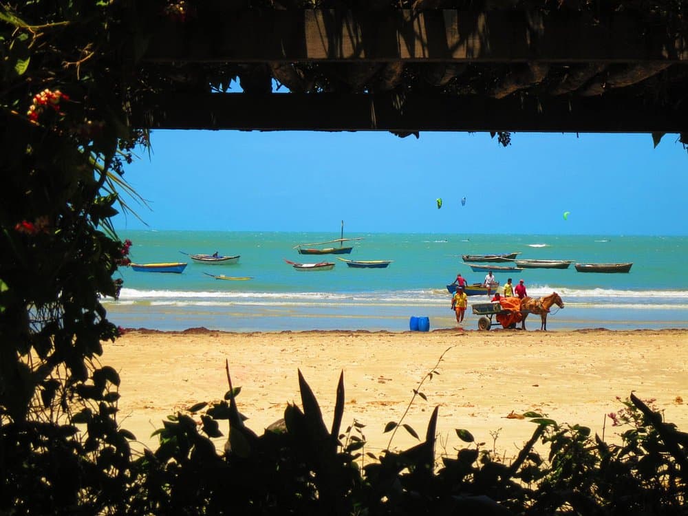 Praia de Mundaú com jangadas ancoradas na água rasa, carroça na areia e kitesuristas ao fundo